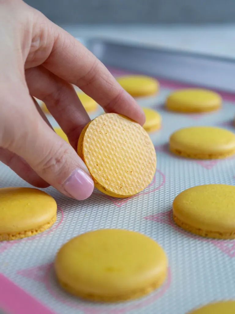 holding up a cooled yellow macaron shell that was properly baked and has a nice shiny, smooth bottom