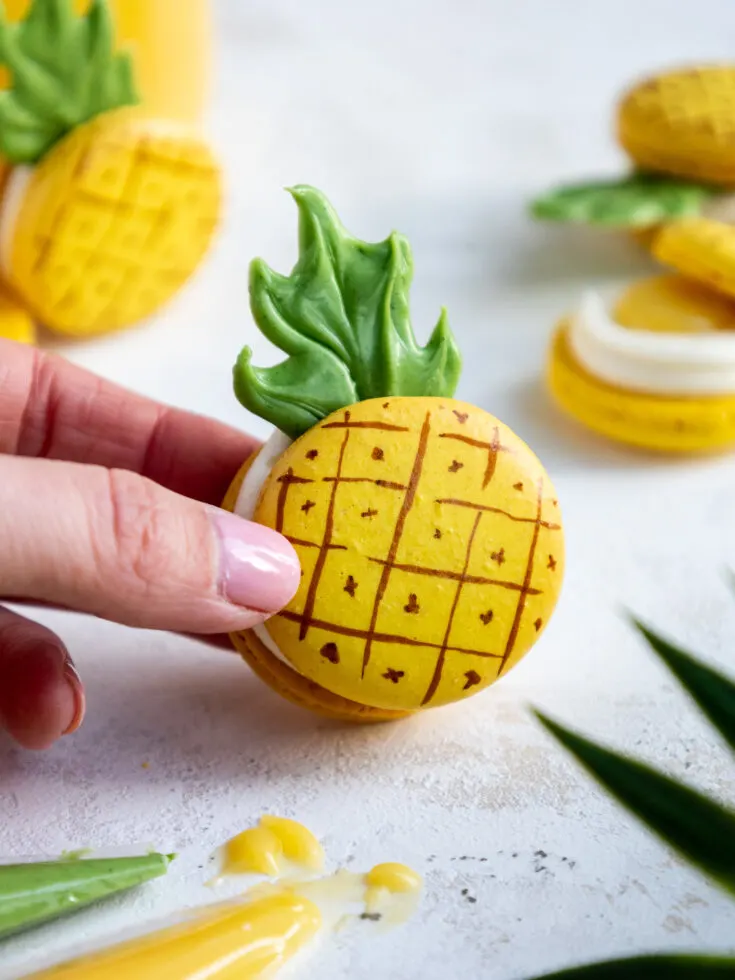 holding up a pineapple shaped macaron that's filled with cream cheese buttercream and pineapple curd