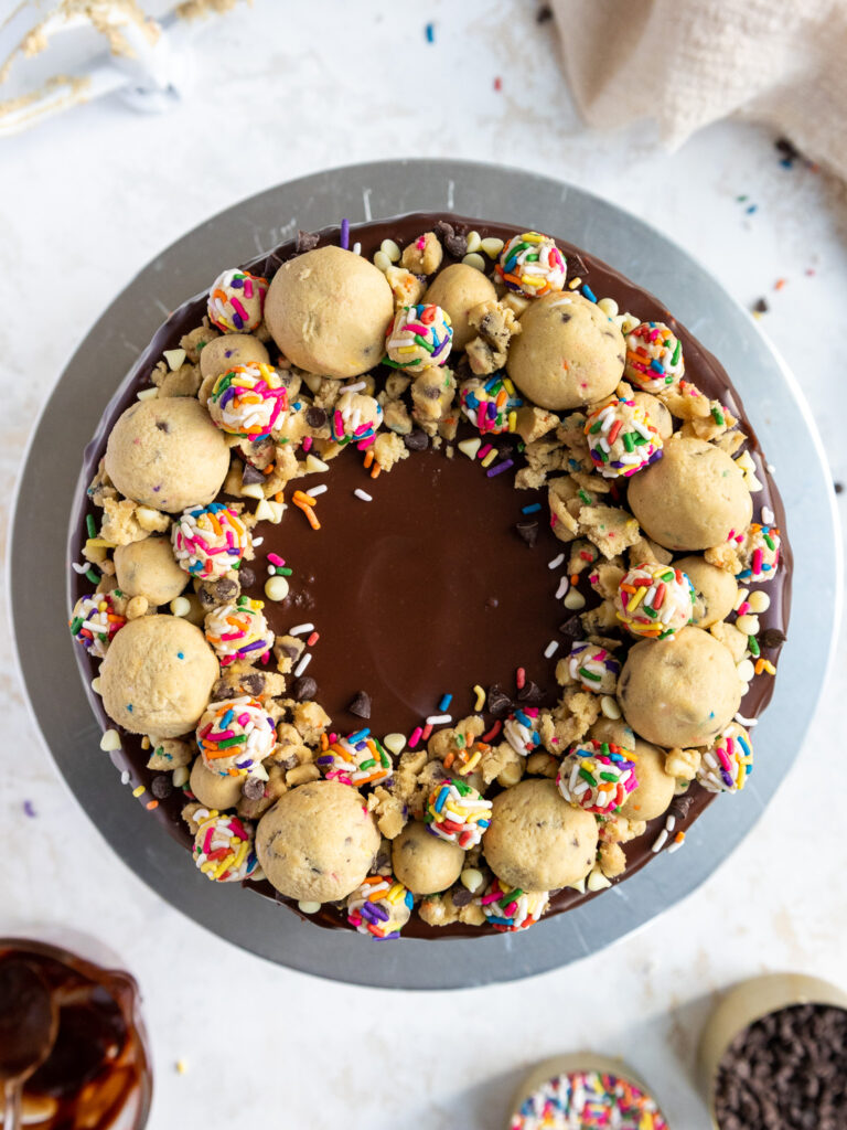 image of a cookie dough cake from overhead, decorated with chocolate ganache and a ring of edible cookie dough balls