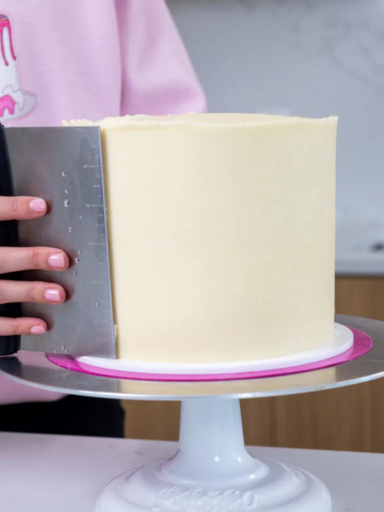 a second layer of cream cheese buttercream being smoothed onto a pink velvet cake with a warm metal bench scraper