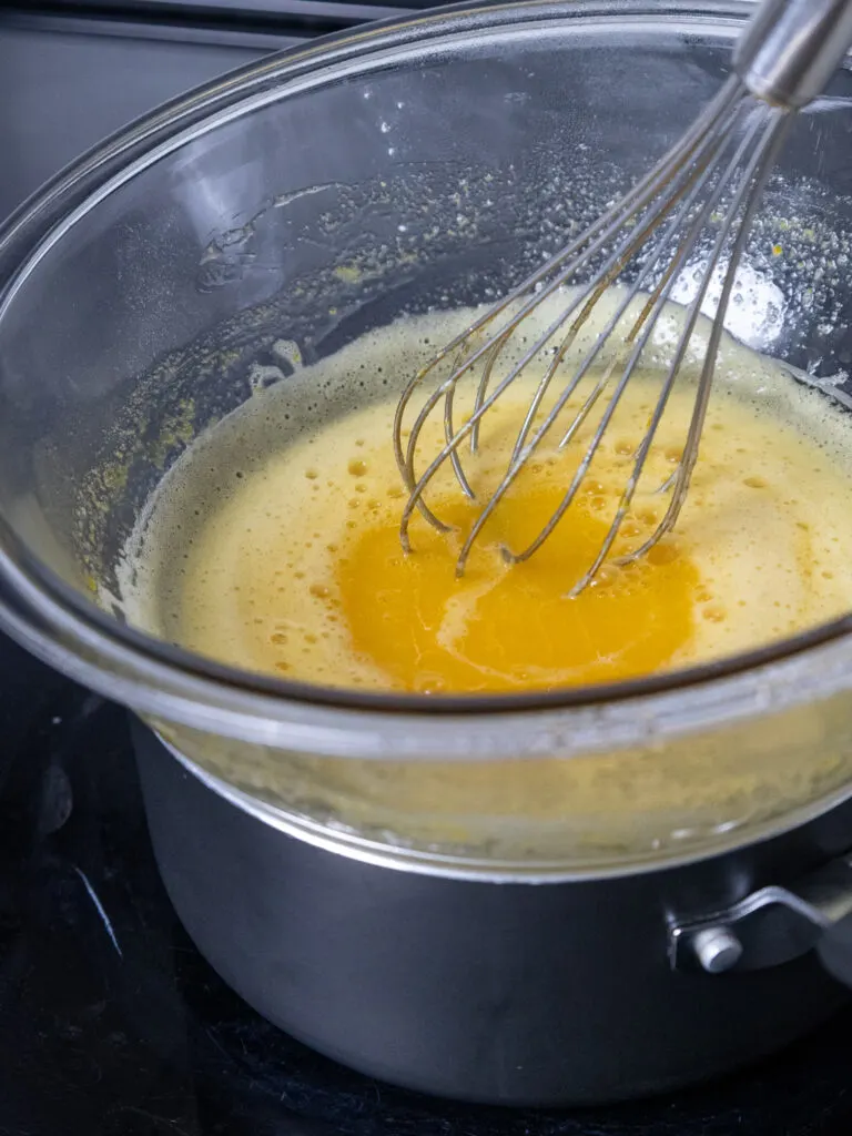 pineapple curd base in a glass bowl on top of a double boiler that is being cooked slowly and whisked continuously