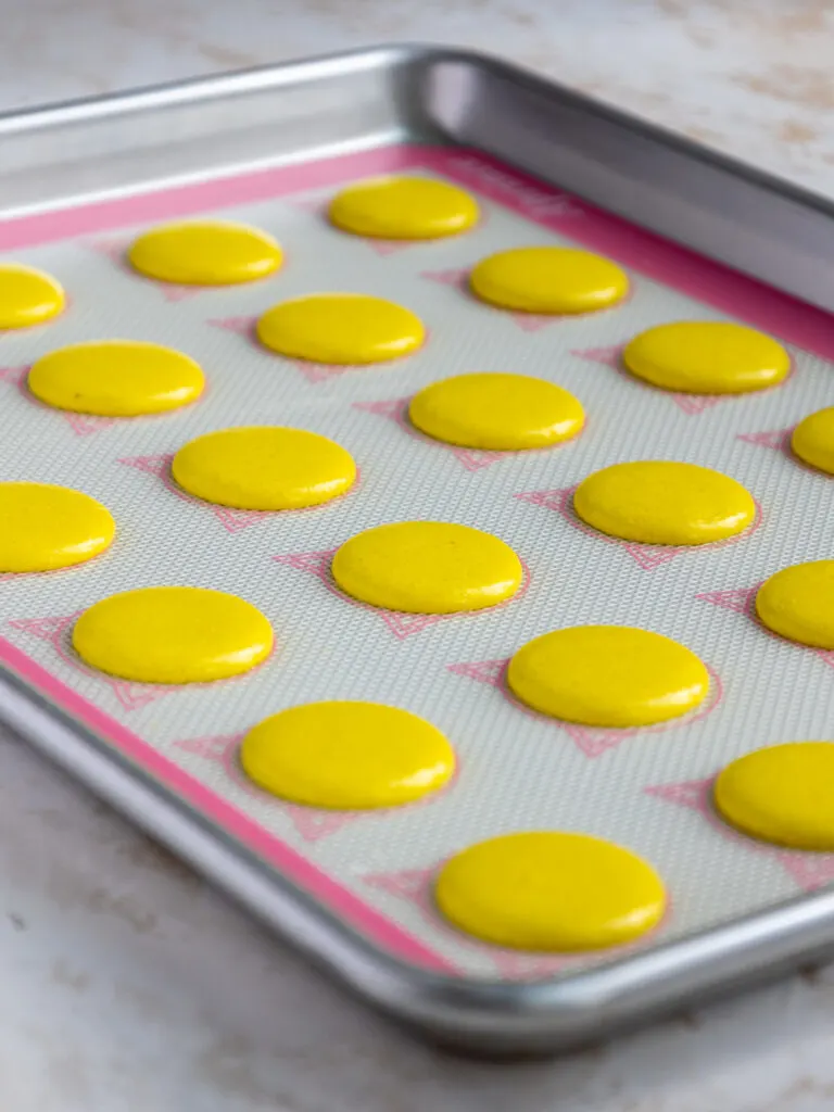 yellow french macaron shells that have been piped on a silpat mat and are resting before being baked