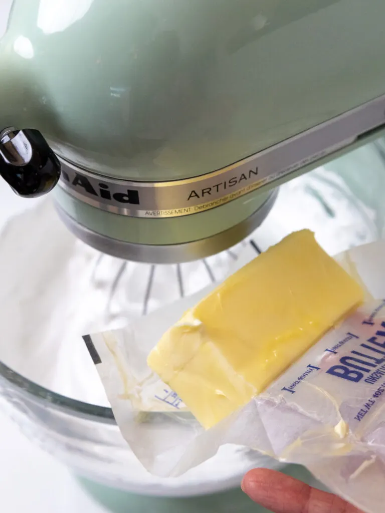 image of a stick of room temperature butter being added into Swiss meringue to make Swiss meringue buttercream