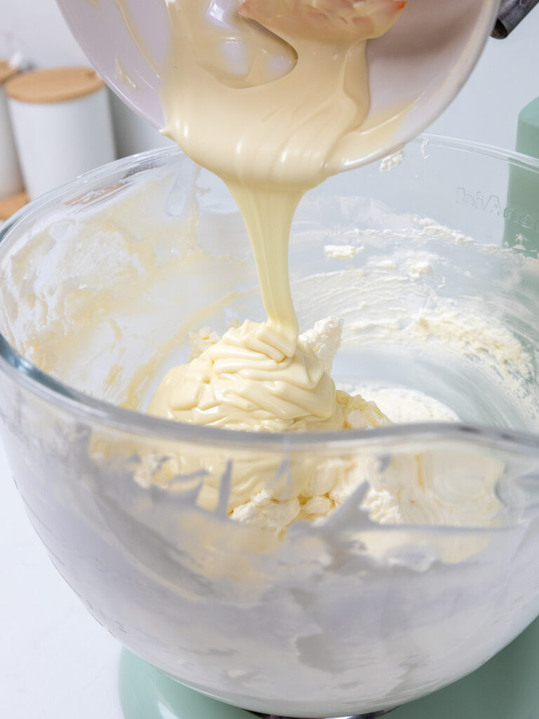 image of melted and cooled white chocolate being poured into Swiss meringue buttercream to make white chocolate Swiss meringue buttercream
