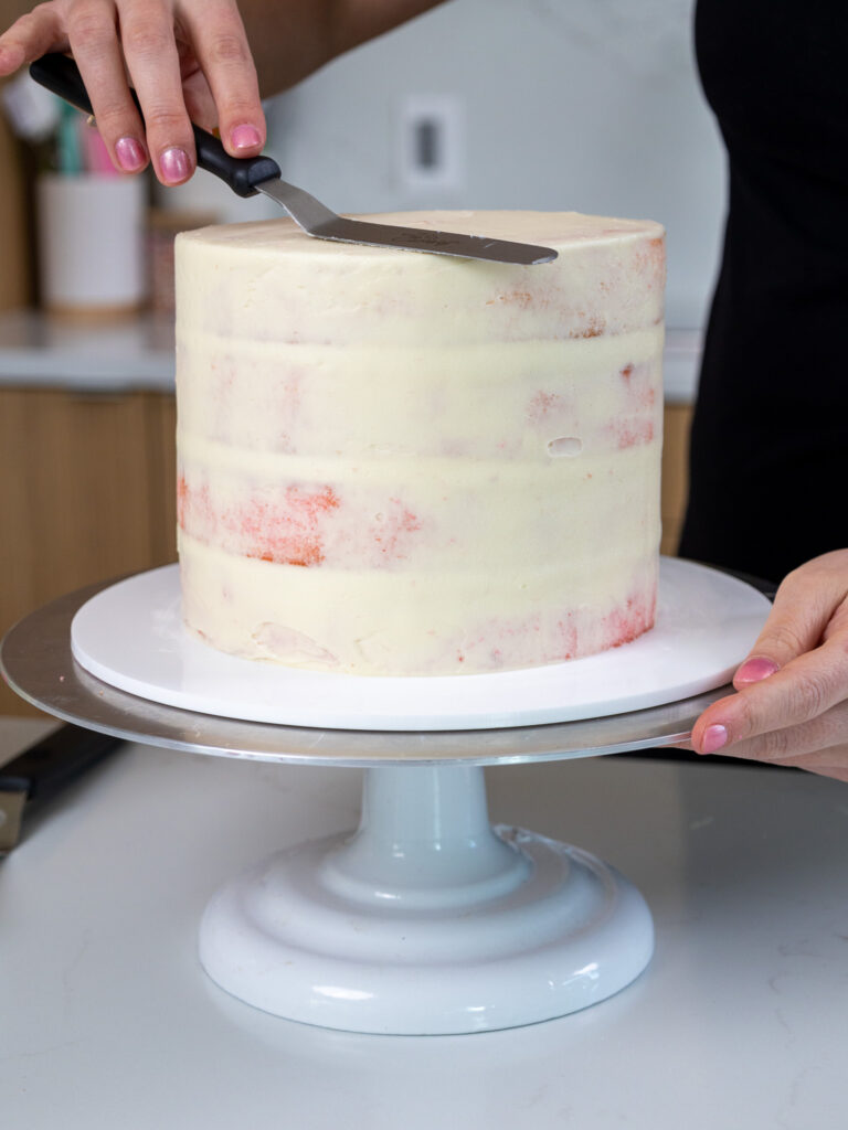 image of pink cake layers being crumb coated with American buttercream and smoothed with small offset spatula