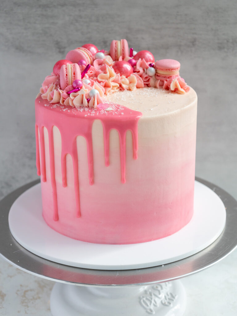 image of a pretty pink cake decorated with ombre pink buttercream, a pink ganache drip, and pink macarons 