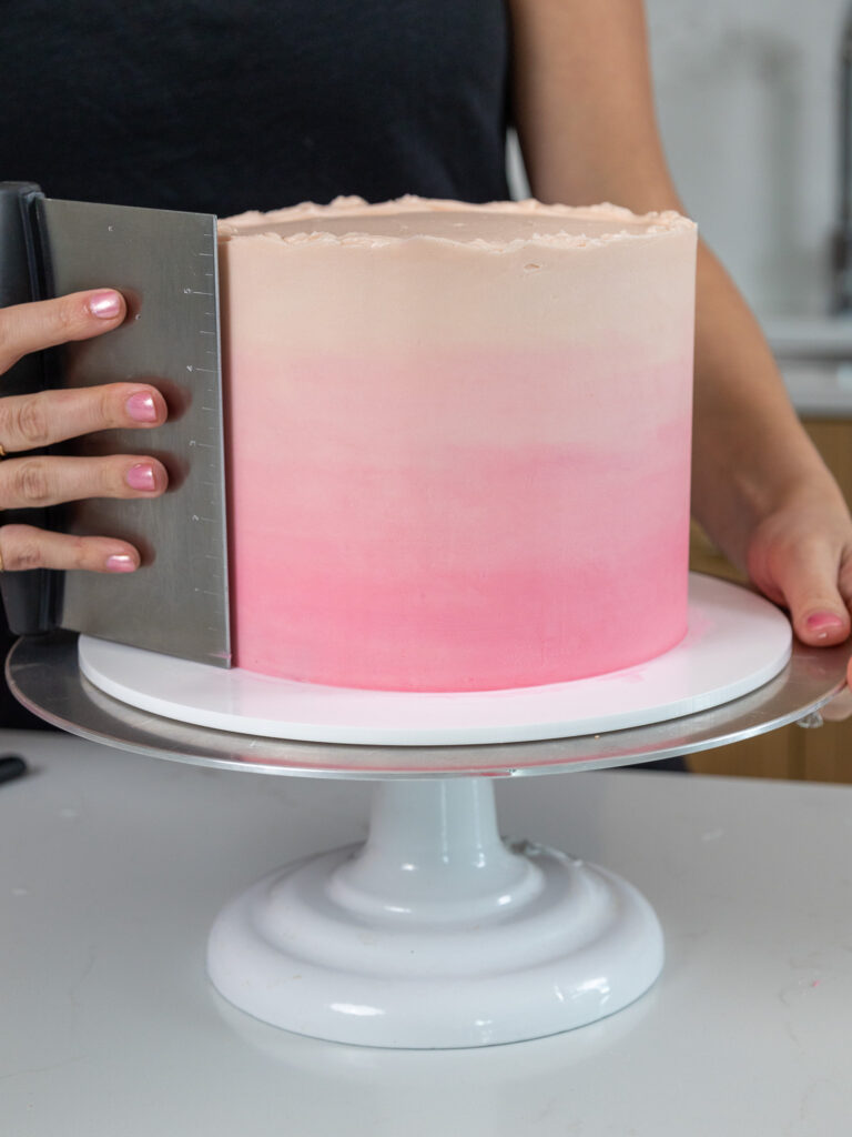 pink ombre frosting being smoothed on a cake with a metal bench scraper