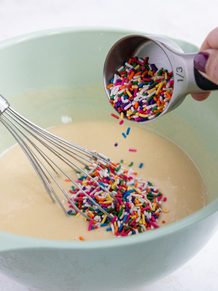 image of rainbow sprinkles being poured into vanilla cupcake batter