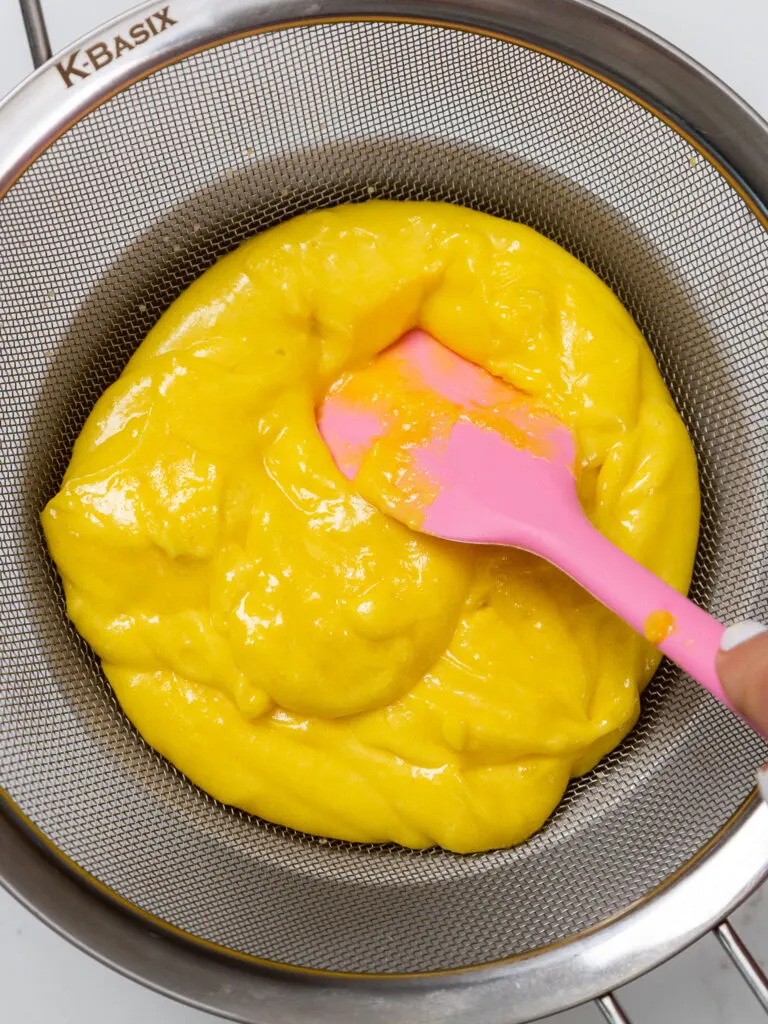image of lemon curd being pushed through a metal strainer to make it smooth