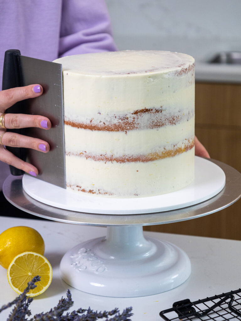image of a lavender lemon cake being crumb coated and having the frosting smoothed with a metal bench scraper