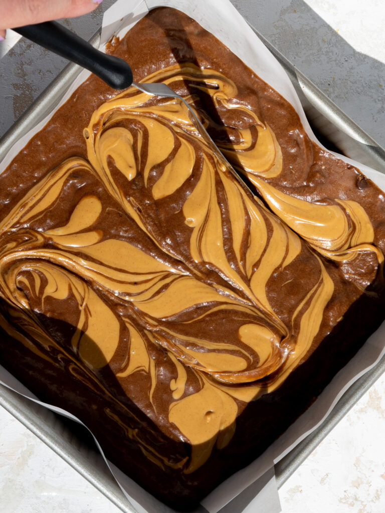 image of warm peanut butter being swirled into banana brownie batter with a small offset spatula