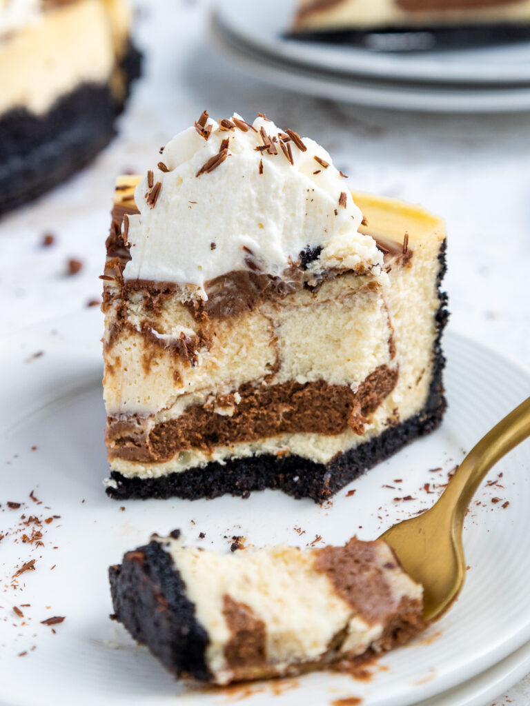 image of a slice of marbled cheesecake on a plate that's been decorated with whipped cream and chocolate shavings and that has been cut into to show its fluffy, light texture