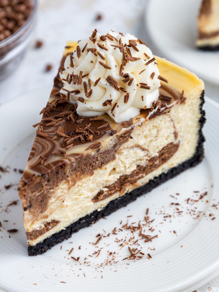 image of a slice of marbled cheesecake on a plate that's been decorated with whipped cream and chocolate shavings