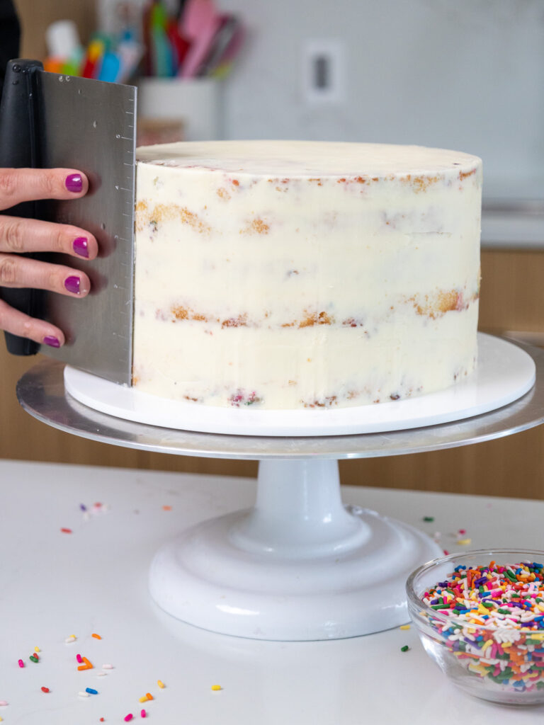 image of a funfetti layer cake being crumb coated and smoothed with a bench scraper