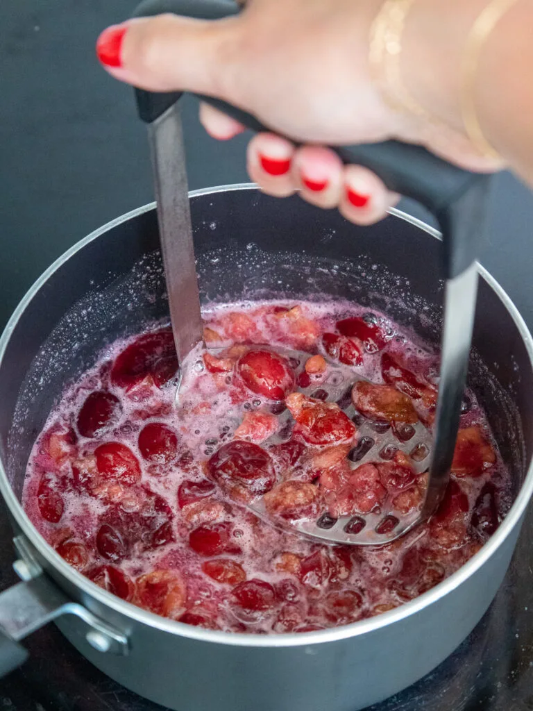 image of cherries being mashed with a potato masher in a saucepan while cooking