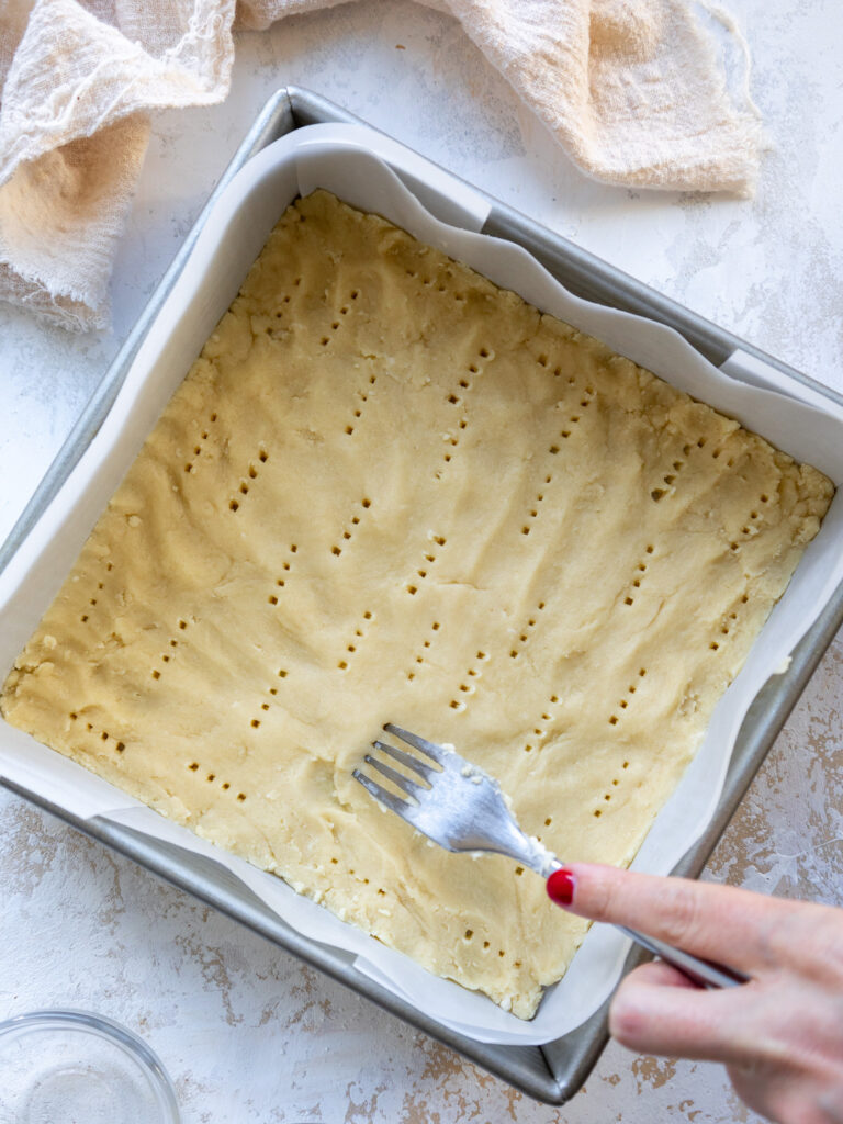 image of shortbread crust being pressed into a square pan and poked with a fork before being baked