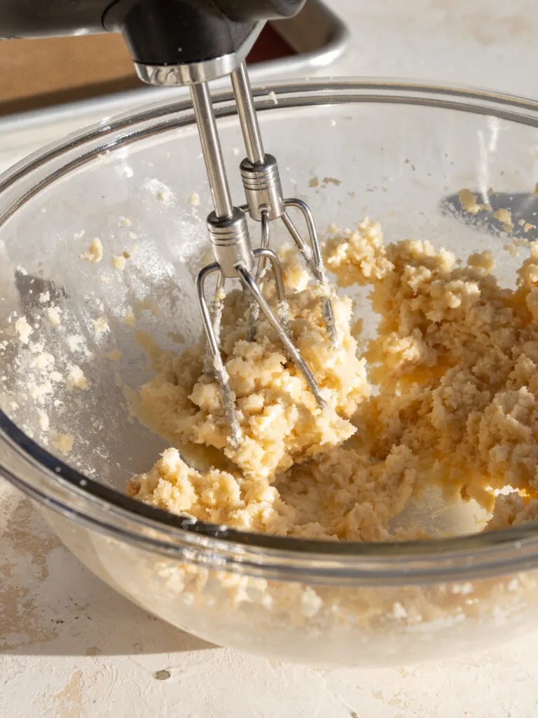 image of butter, brown sugar, and granulated sugar being creamed together in a glass bowl with an electric hand mixer