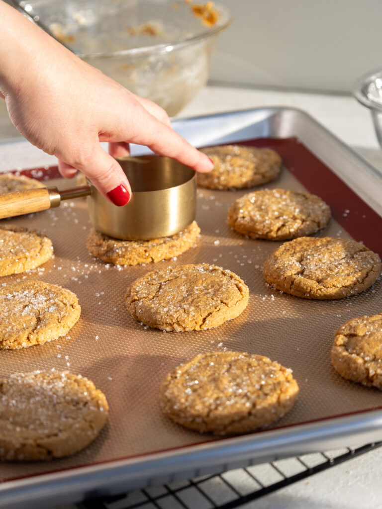 image of baked gingerbread cookies being flattened with the bottom of a metal measuring cup