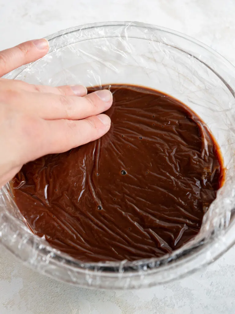 image of dark chocolate ganache macaron filling being covered with saran wrap to prevent a skin from forming before being set aside to cool to room temperature and thicken