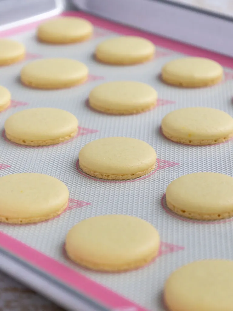 image of baked, light yellow French macaron shells that are cooling on a Silpat mat and that have perfect feet