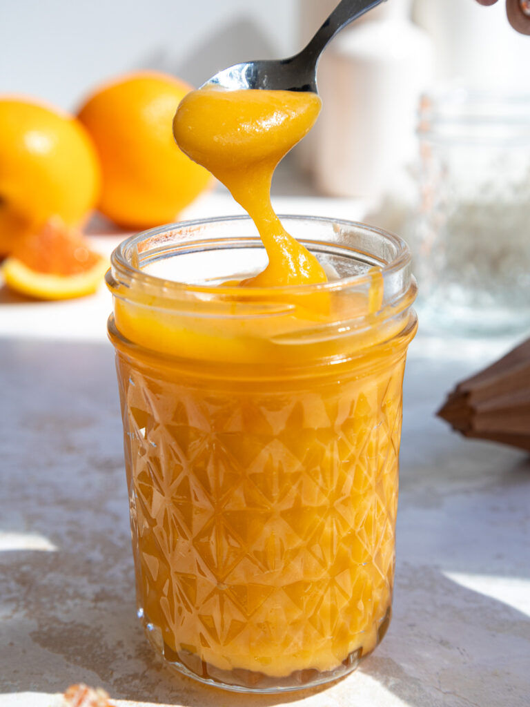 image of orange curd that's being scooped out of a glass container with a small metal spoon