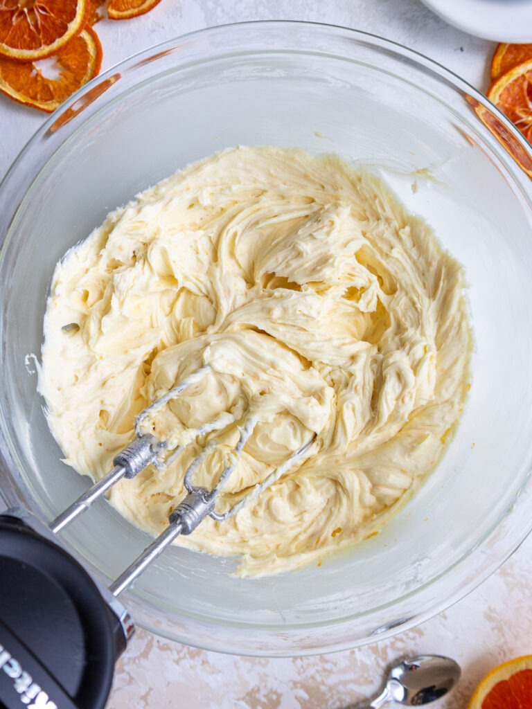 image of orange buttercream being made in a glass bowl with an electric hand mixer