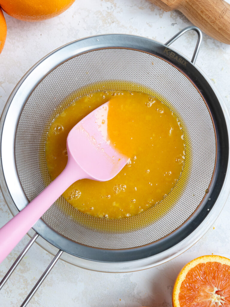 image of orange curd being poured through a fine mesh strainer to remove any bits of cooked egg, orange pulp, and orange seeds.