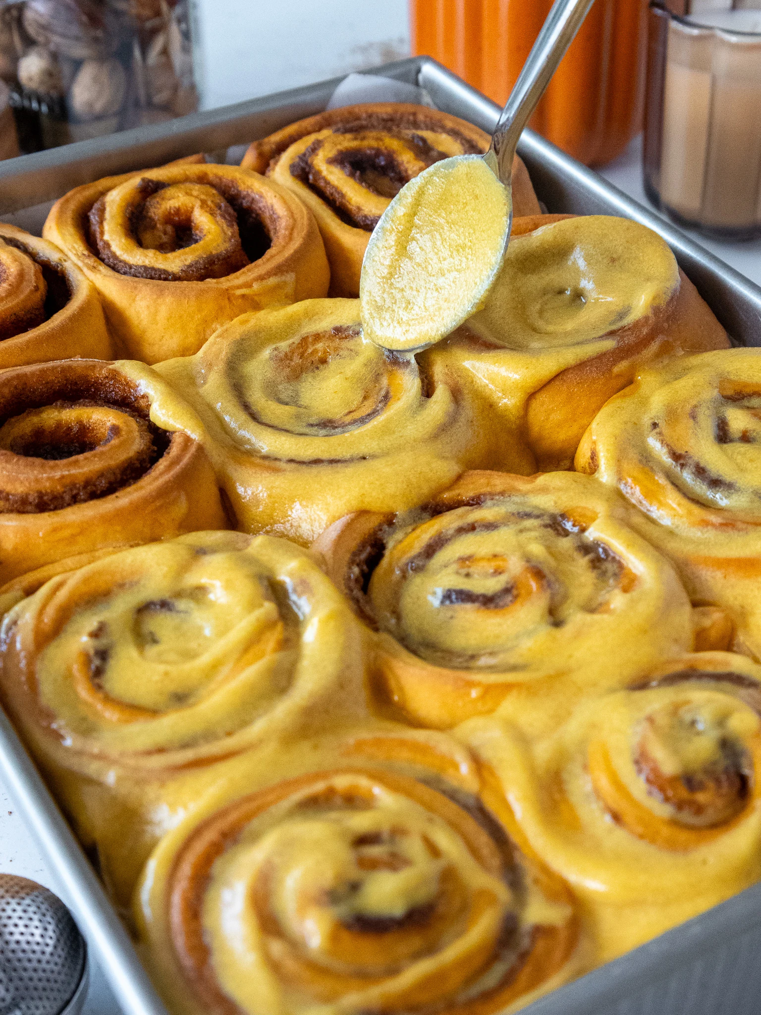 image of pumpkin pie cinnamon rolls being frosted with pumpkin cream cheese frosting