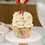 image of not too sweet buttercream that's been piped onto a cupcake and is being topped with rainbow sprinkles