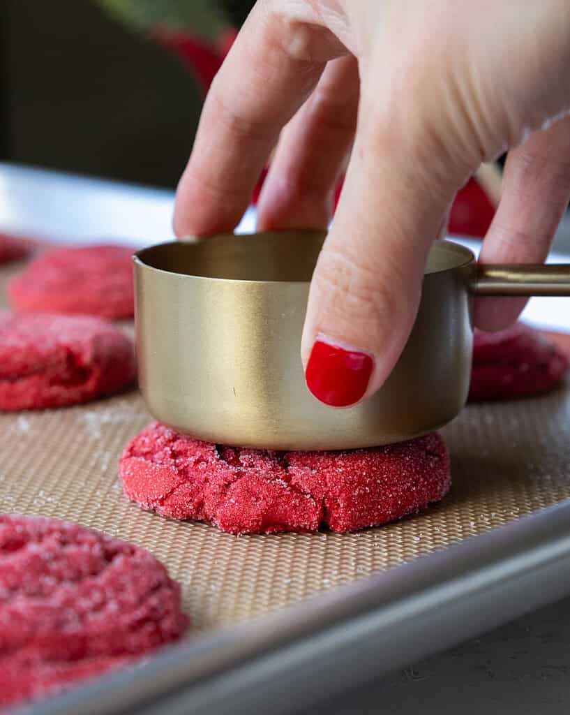 image of a red velvet sugar cookie being flattened out with a measuring up to give it a perfectly chewy texture