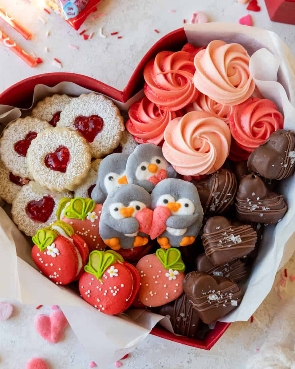 image of chelsweets 2026 valentine's day cookie boxes, made with raspberry linzers, strawberry macarons, rose meringues, chocolate peanut butter hearts, and penguin marshmallows